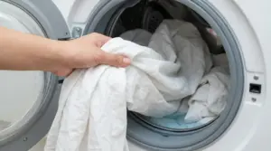 Una mano inserisce lenzuola bianche in una lavatrice a carica frontale